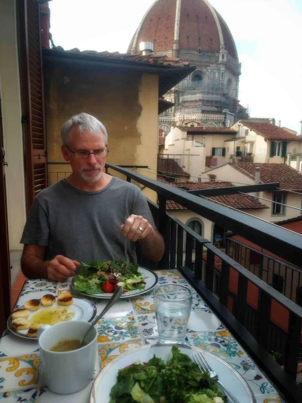 Man with grey hair eating a salad on a small balcony with the Florence Duomo dome in the background.