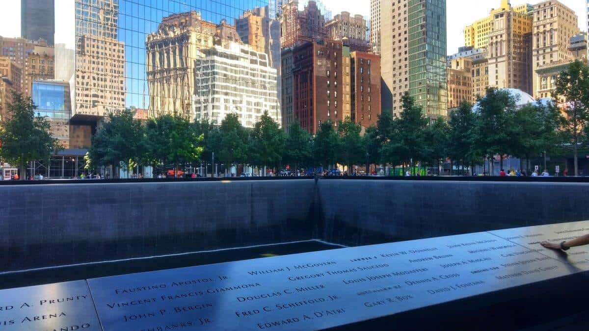 9/11 Memorial pool in New York City showing names engraved in bronze with buildings and trees in the background.