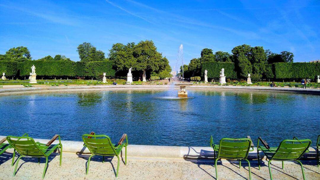 Green metal chairs around a large fountain basin in a park in Paris.