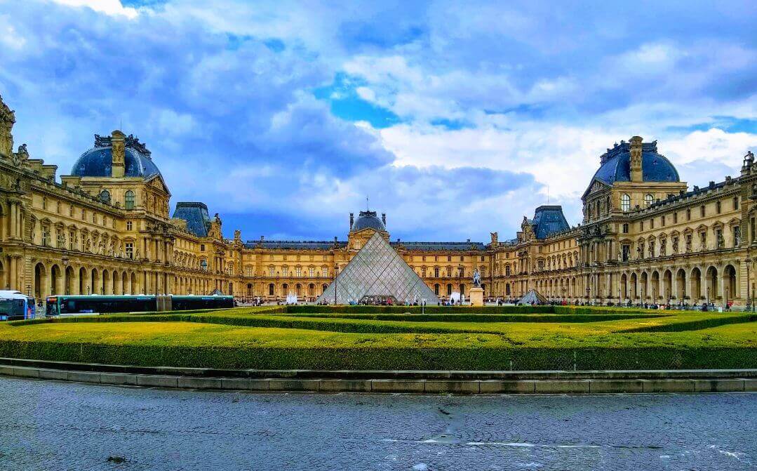 The Louvre pyramid in front of the Louvre palace with a blue sky.