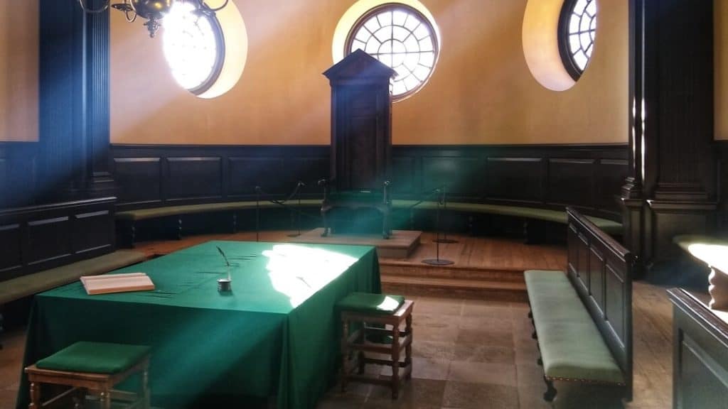 Large table with a green table cloth in a room with wooden benches and sun streaming through round windows.