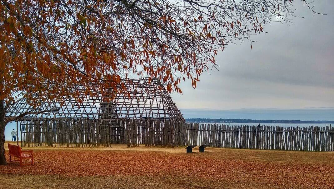 Frame wooden building by an old wooden fence on a river with leaves on the ground.