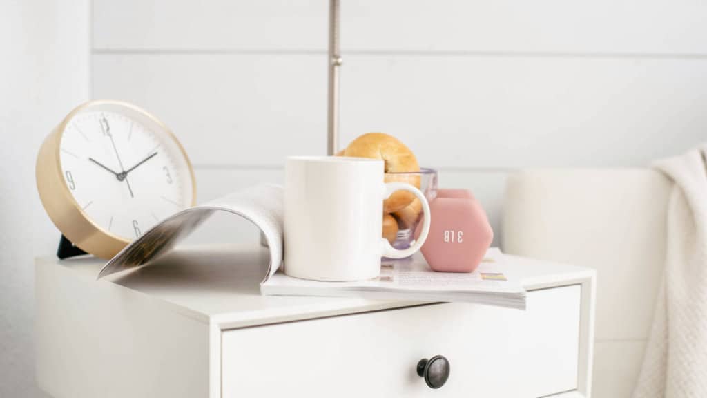 White coffee cup, magazine, bagels and a clock on a white nightstand
