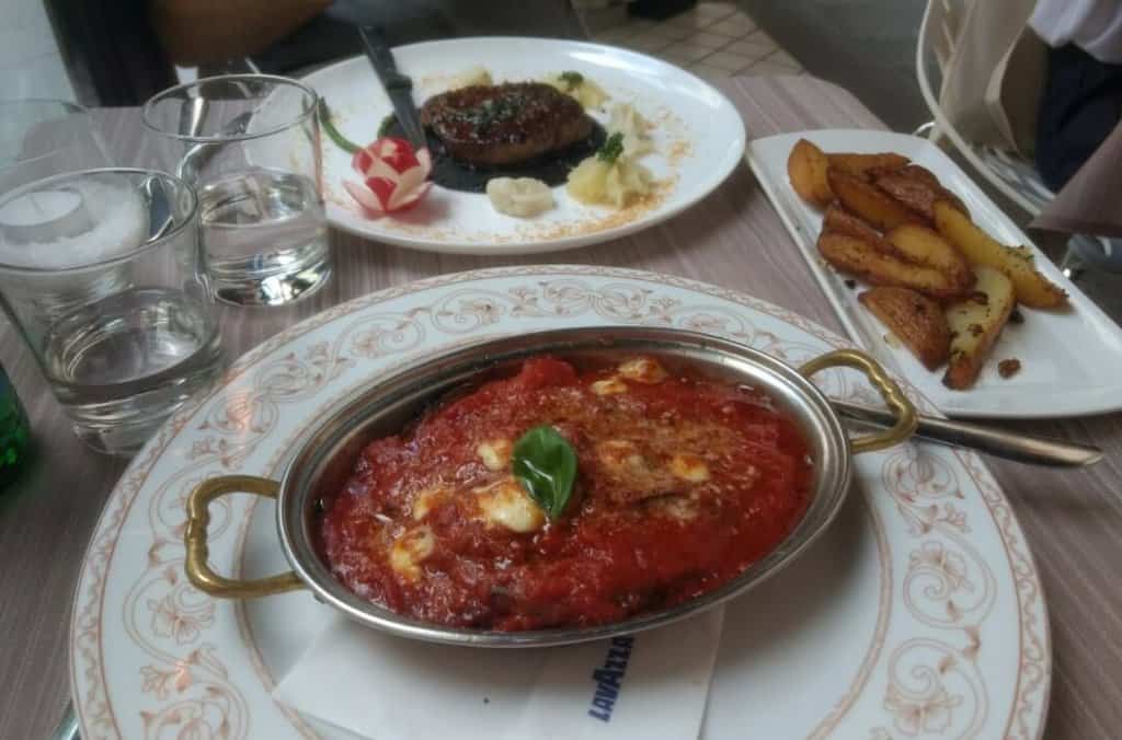 Eggplant parmesan in an oval metal dish alongside a plate of potatoes and a steak