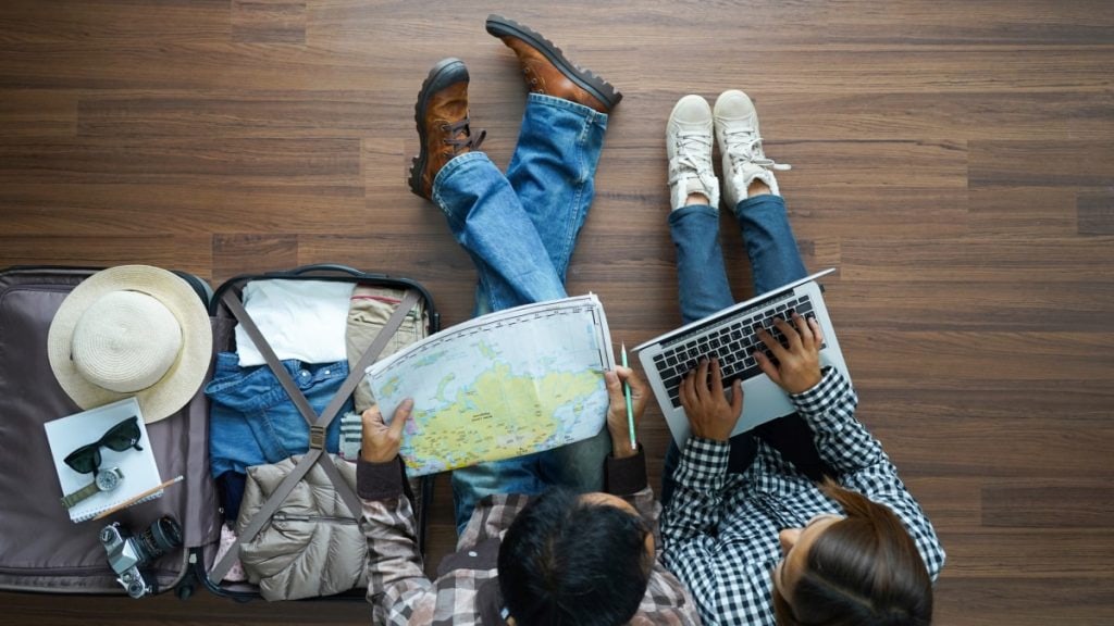 overhead view of a couple sitting on a dark wood floor planning a trip using a paper map and a laptop