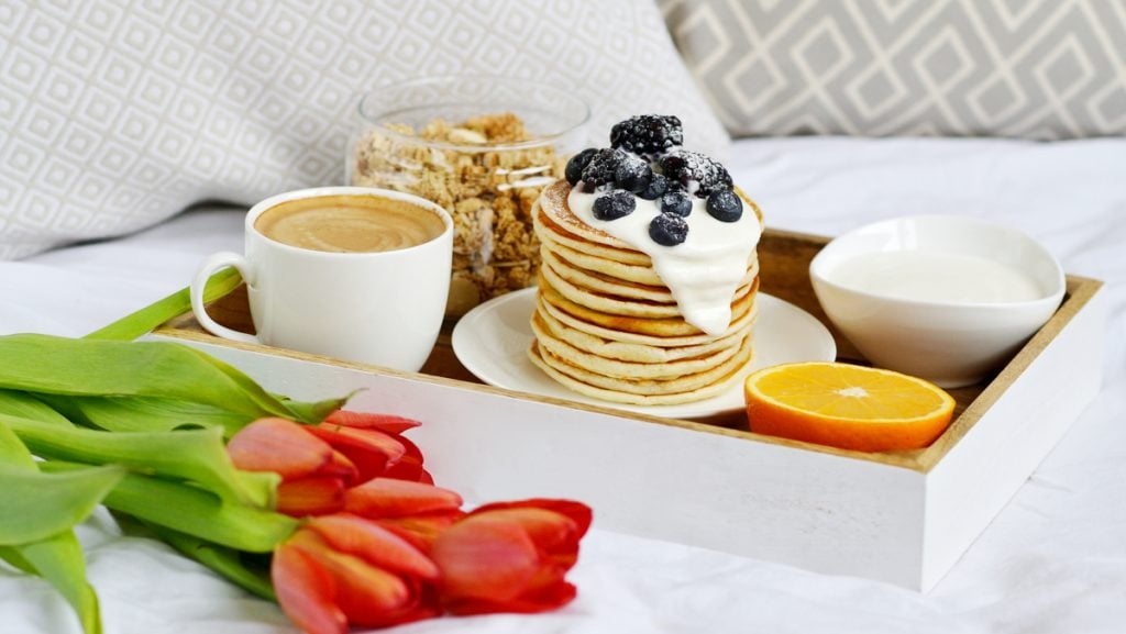 A plate of pancakes, a bowl of granola and a cappuccino on a white tray on a bed with red tulips
