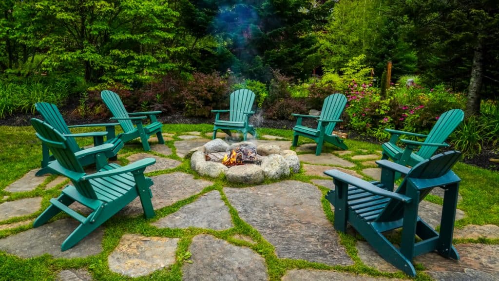 Seven green Adirondack chairs around a stone firepit in a wooded garden area