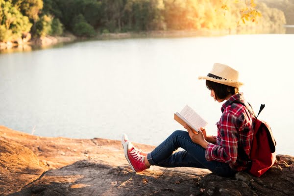 woman wearing a red backback sitting by a lake and reading a book
