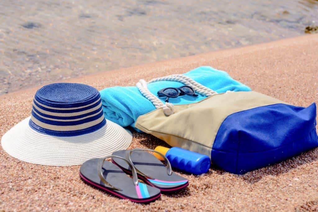 Assorted beach accessories on the sand of a tropical beach overlooking the water with a sunhat, sunscreen, towel, thongs, sunglasses and beach bag