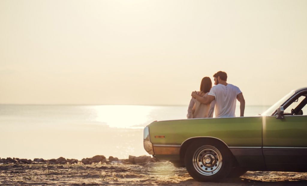 Romantic couple is standing near green retro car on the beach.