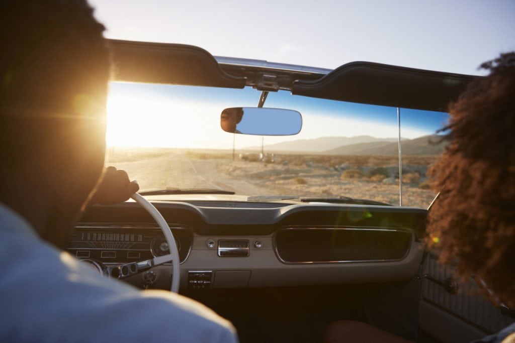 Rear View Of Couple On Road Trip Driving Classic Convertible Car Towards Sunset