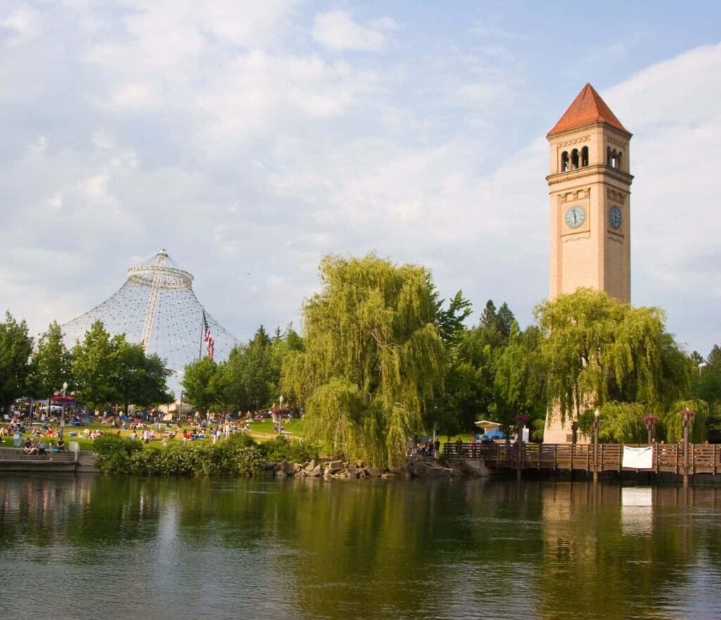 many people in a park on a river with a large clock tower nearby