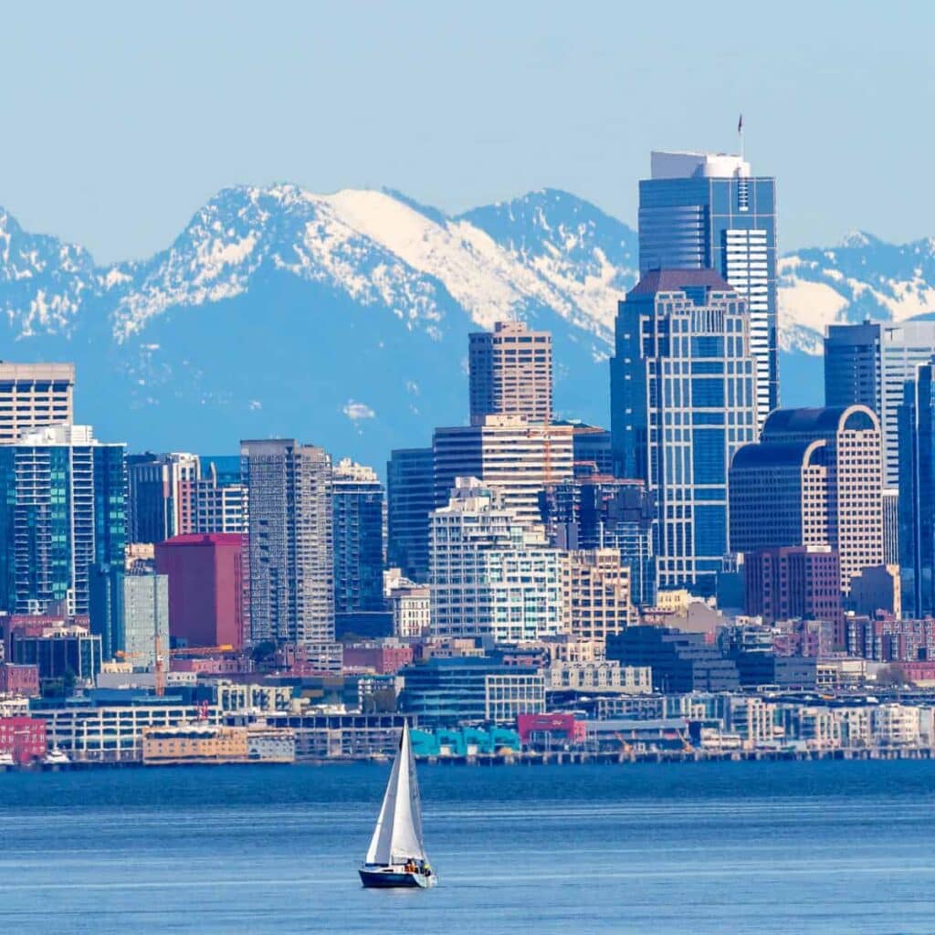 Sailboat on blue water in front of a large city skyline with snow capped mountains in the background