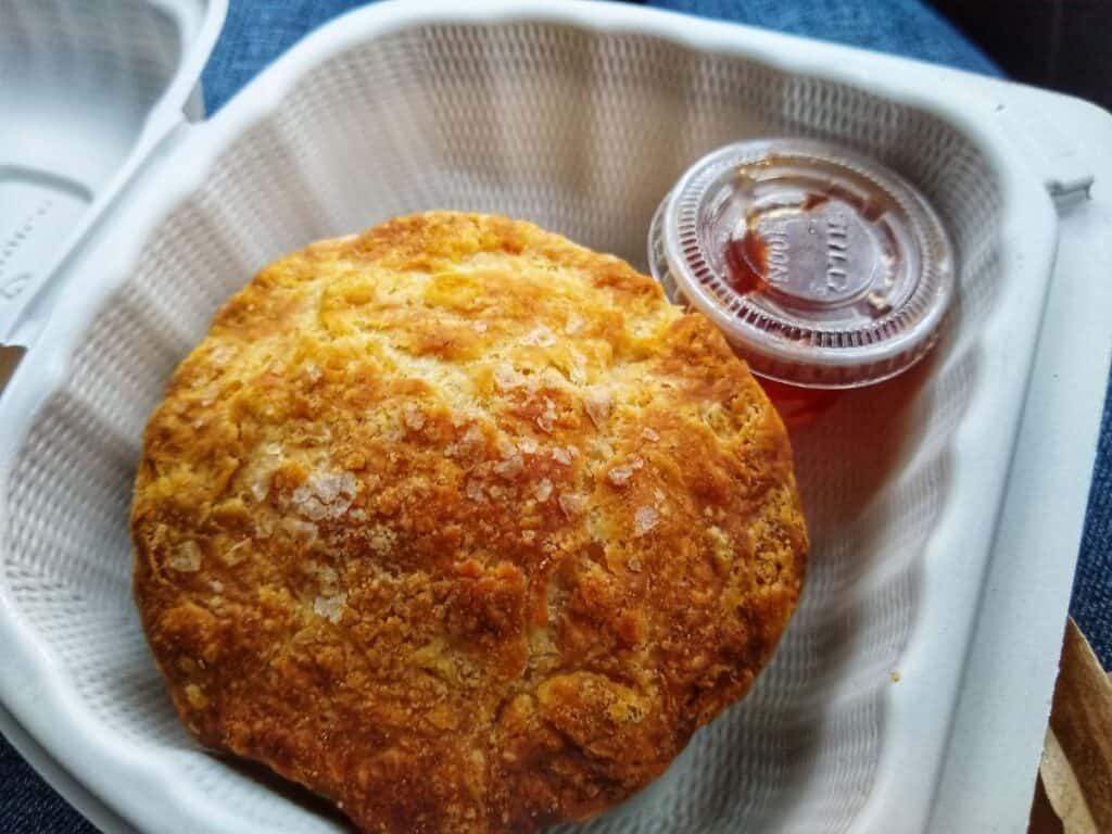 large golden brown biscuit in a takeout container with a small plastic cup of red jam