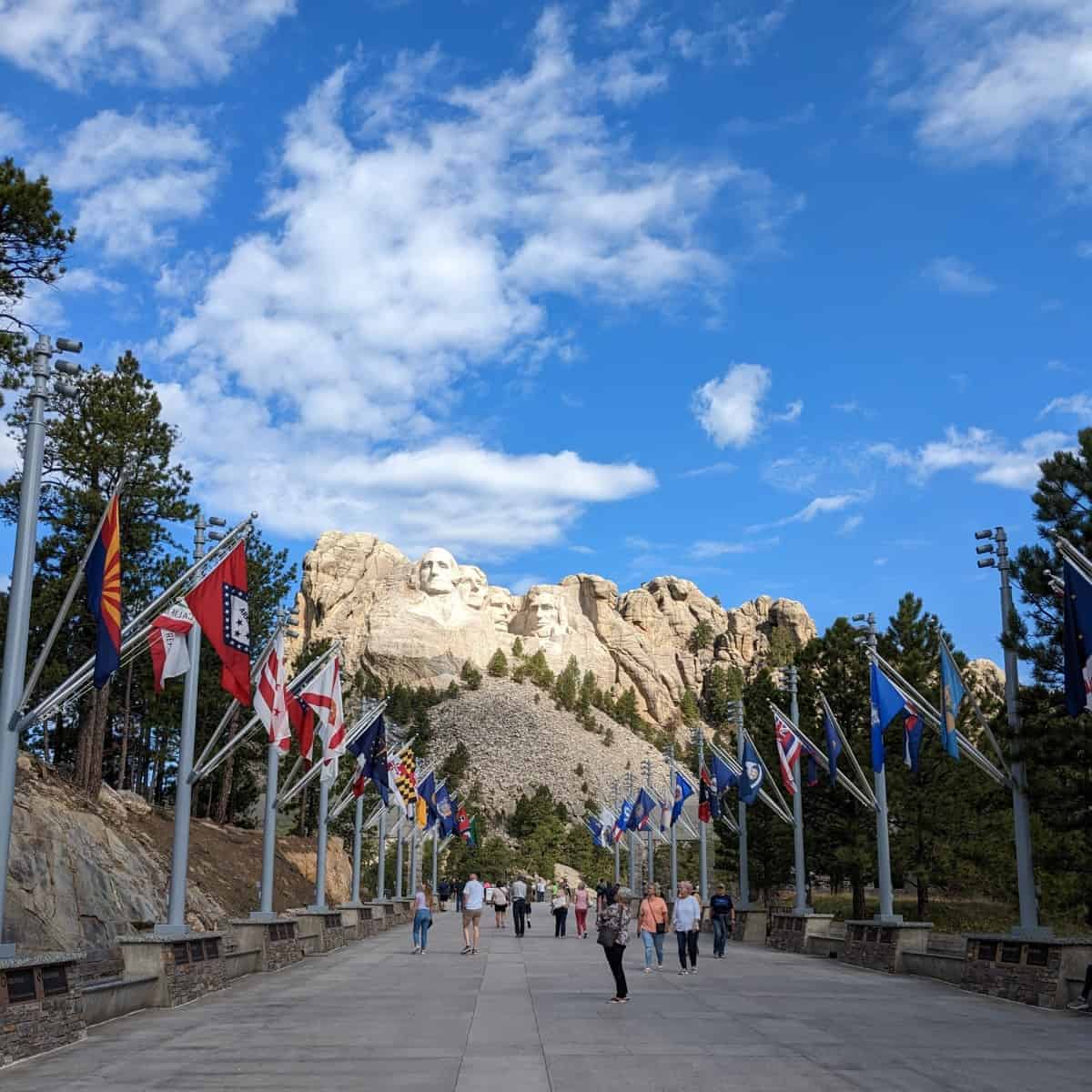 Tourists on a large walkway flanked in state flags, leading to Mount Rushmore.