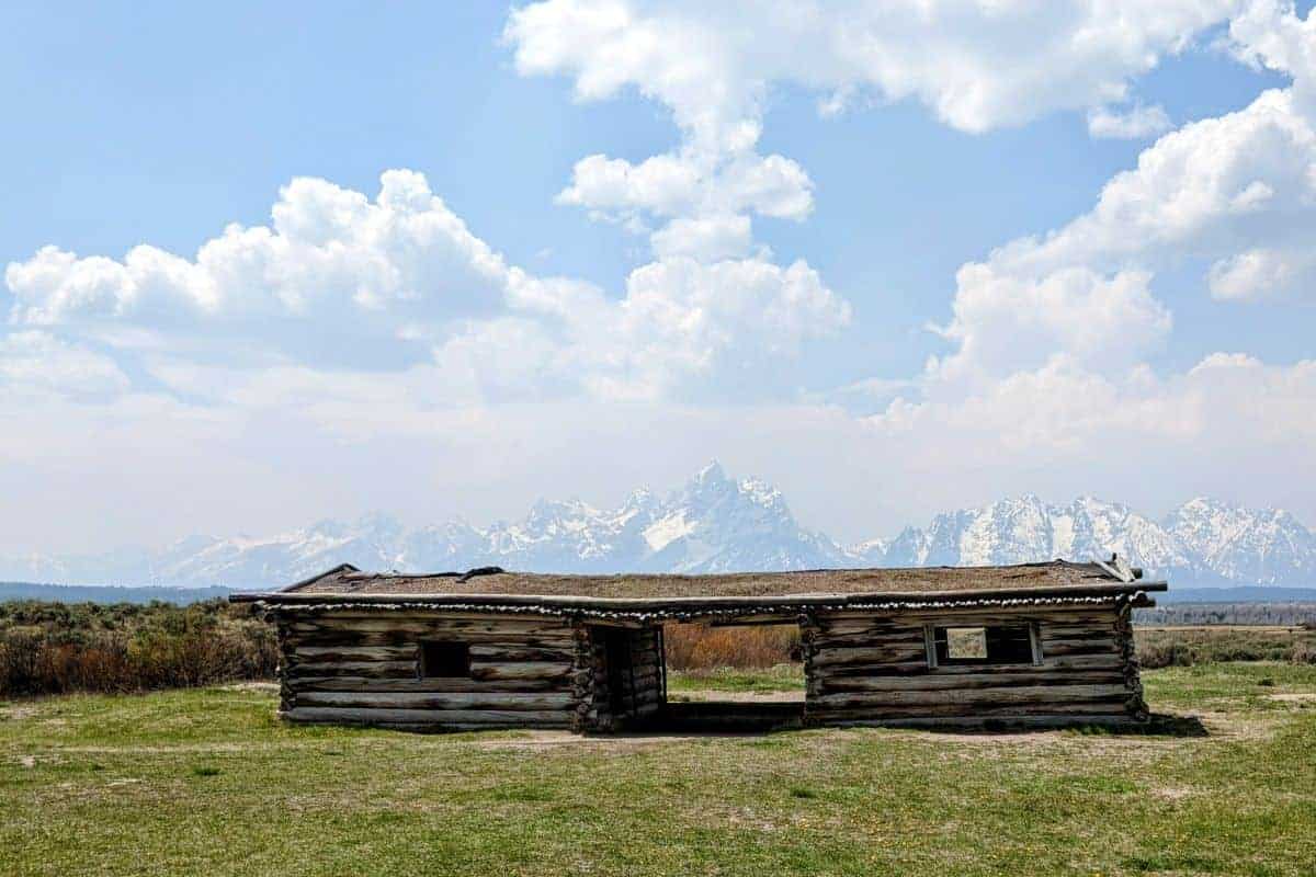 old, rustic cabin surrounded by grass with the Tetons in the background