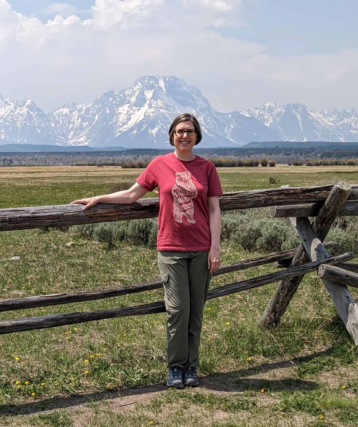woman in hiking gear standing in front of a split rail fence with Mount Moran in the background