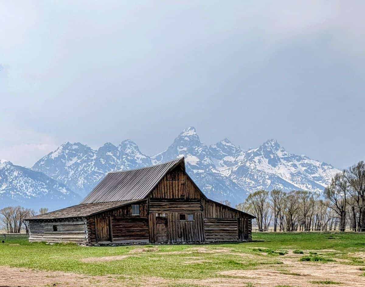 rustic barn with peaked roof surrounded by grass with mountains in the background