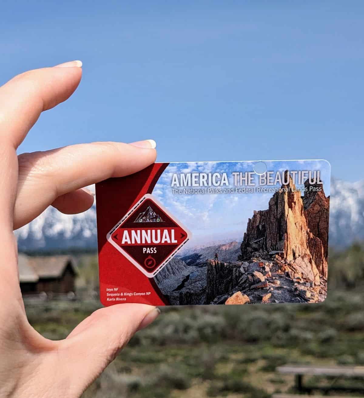Woman's hand holding an America the Beautiful Annual pass with mountains in the background. The pass is one of the best gifts for national park lovers.