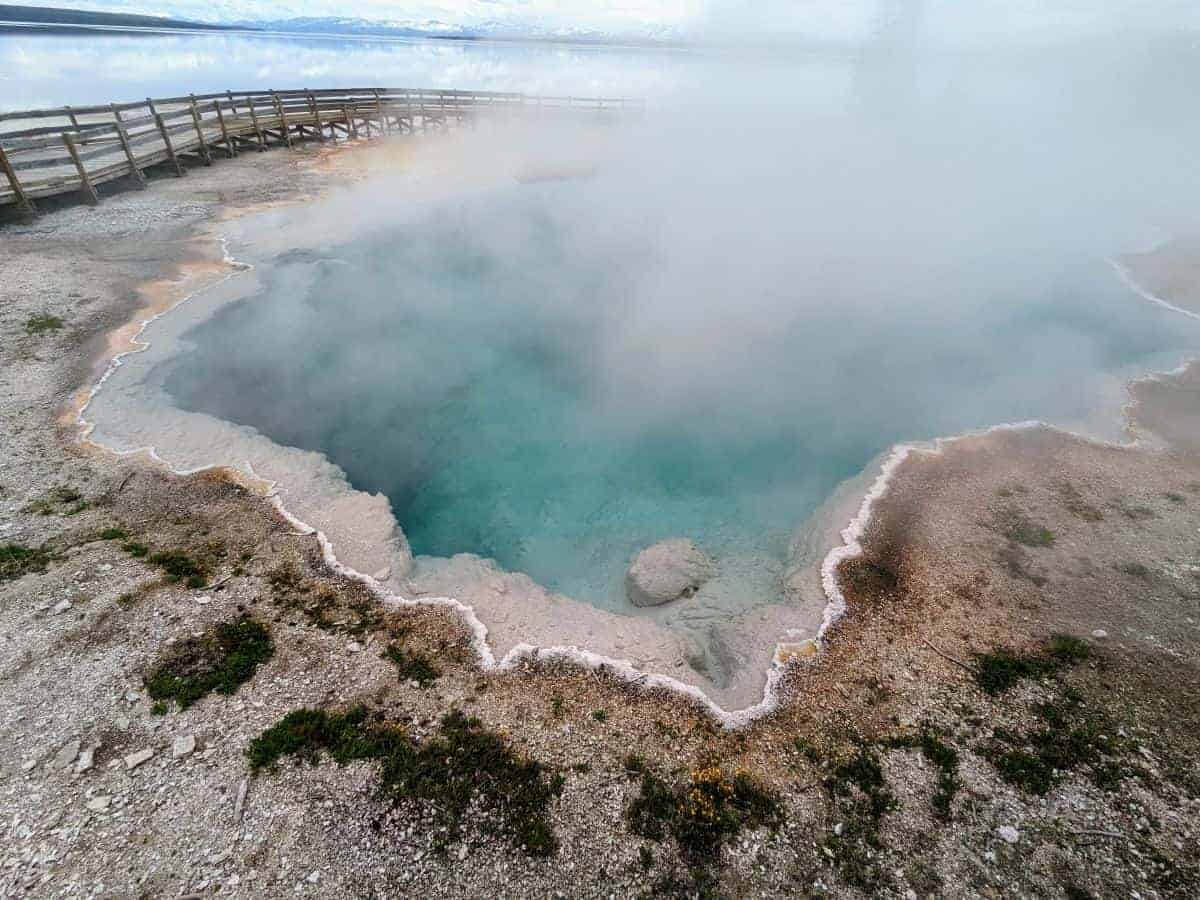 Steam rising off a large turquoise pool. In the background there is a wooden boardwalk with railing. Beyond that there is a large lake with mountains in the far distance.