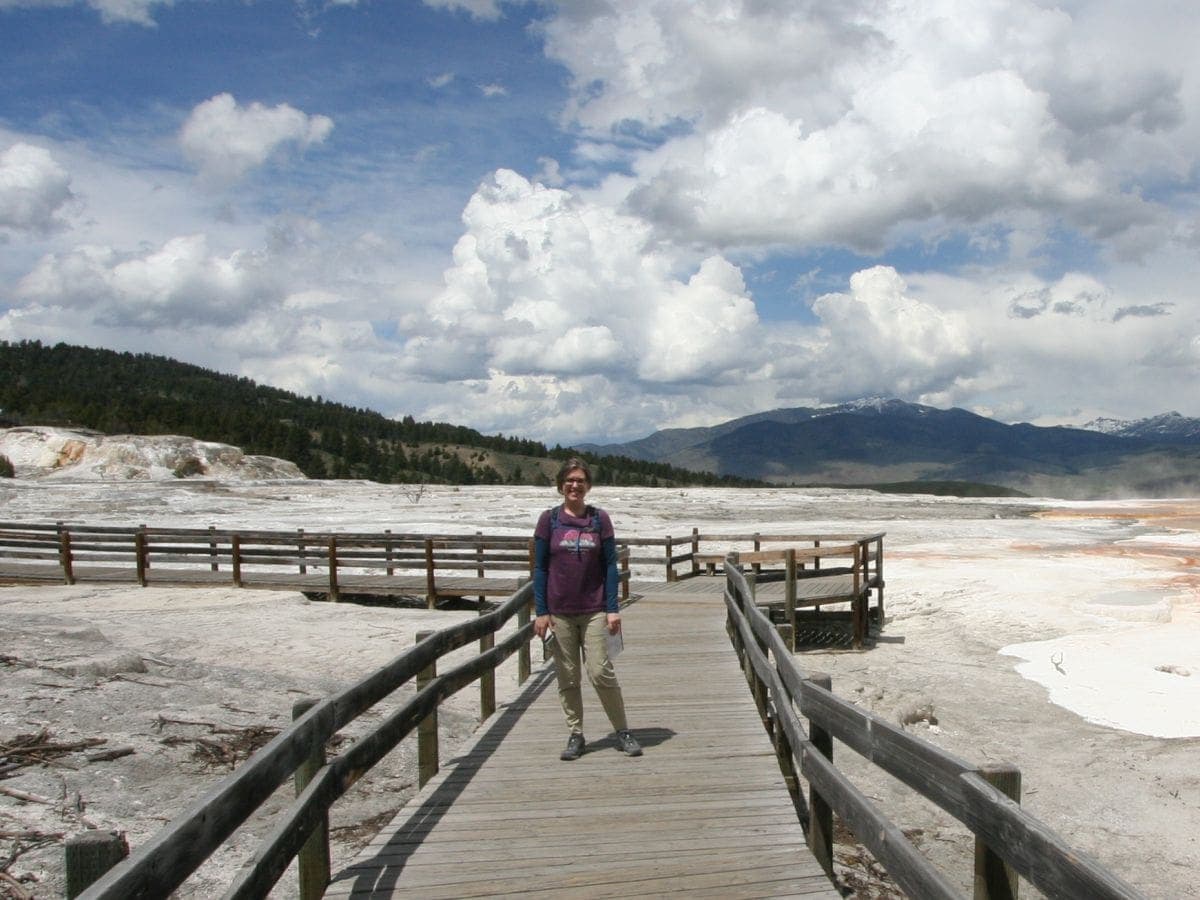 Woman standing on a wooden boardwalk with railings. She is wearing jogger-style hiking pants, hiking shoes, a short-sleeve T-shirt over a long-sleeve base layer and a day hiking backpack.