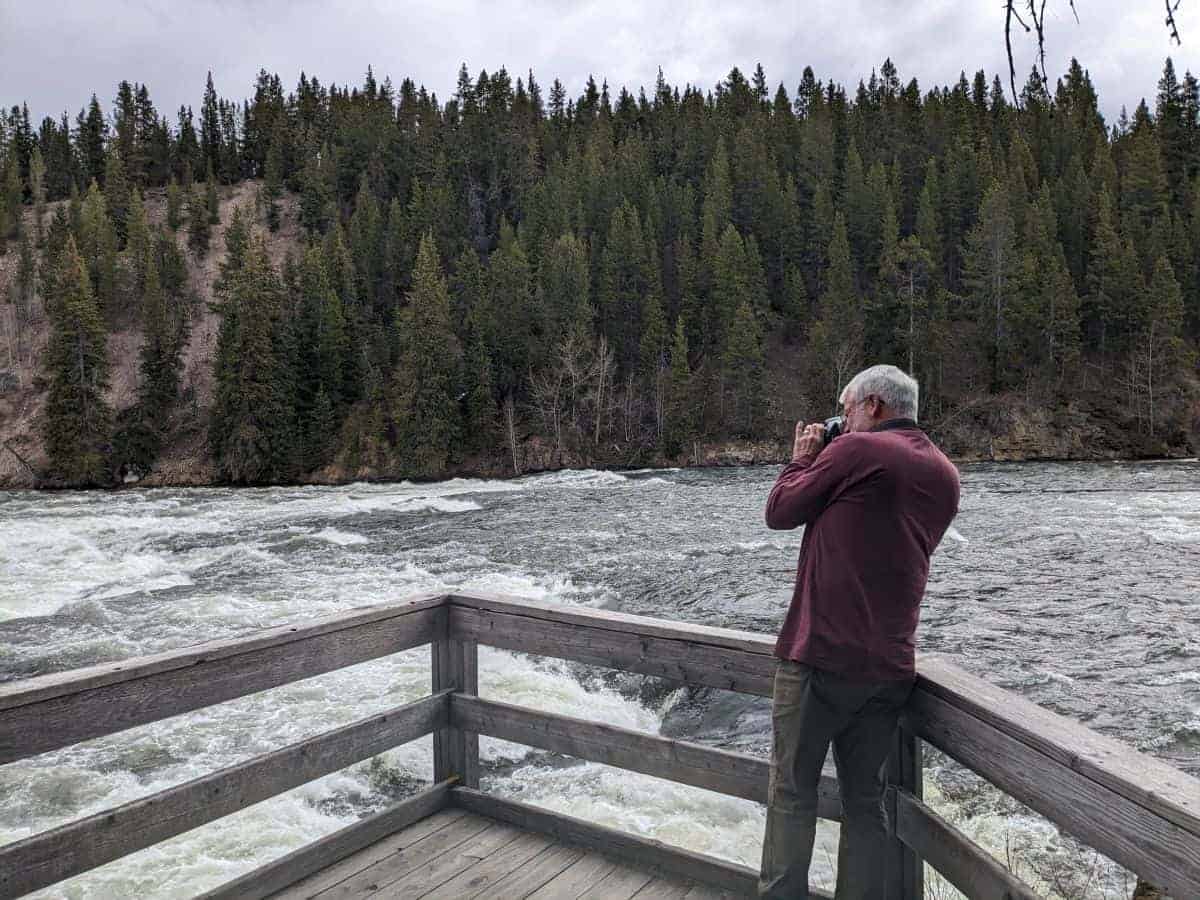 White-haired man holding a DSLR camera and focusing the lens. He is standing at a wooden railing at the edge of some river rapids.