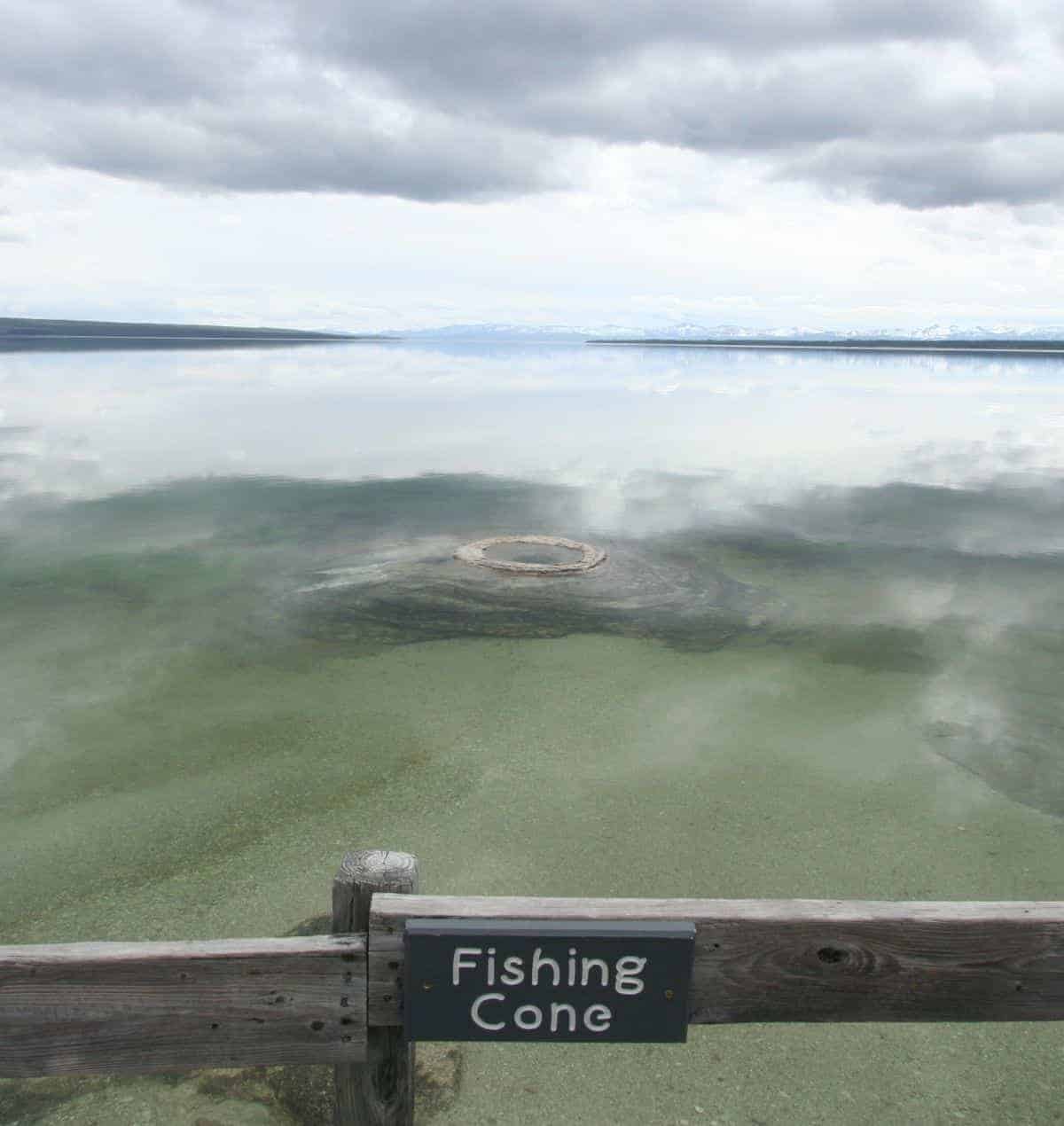 Geyser cone under the water at the edge of a lake. A sign on a wooden railing in the foreground says "Fishing Cone." There are snowy mountains in the distance beyond the lake.