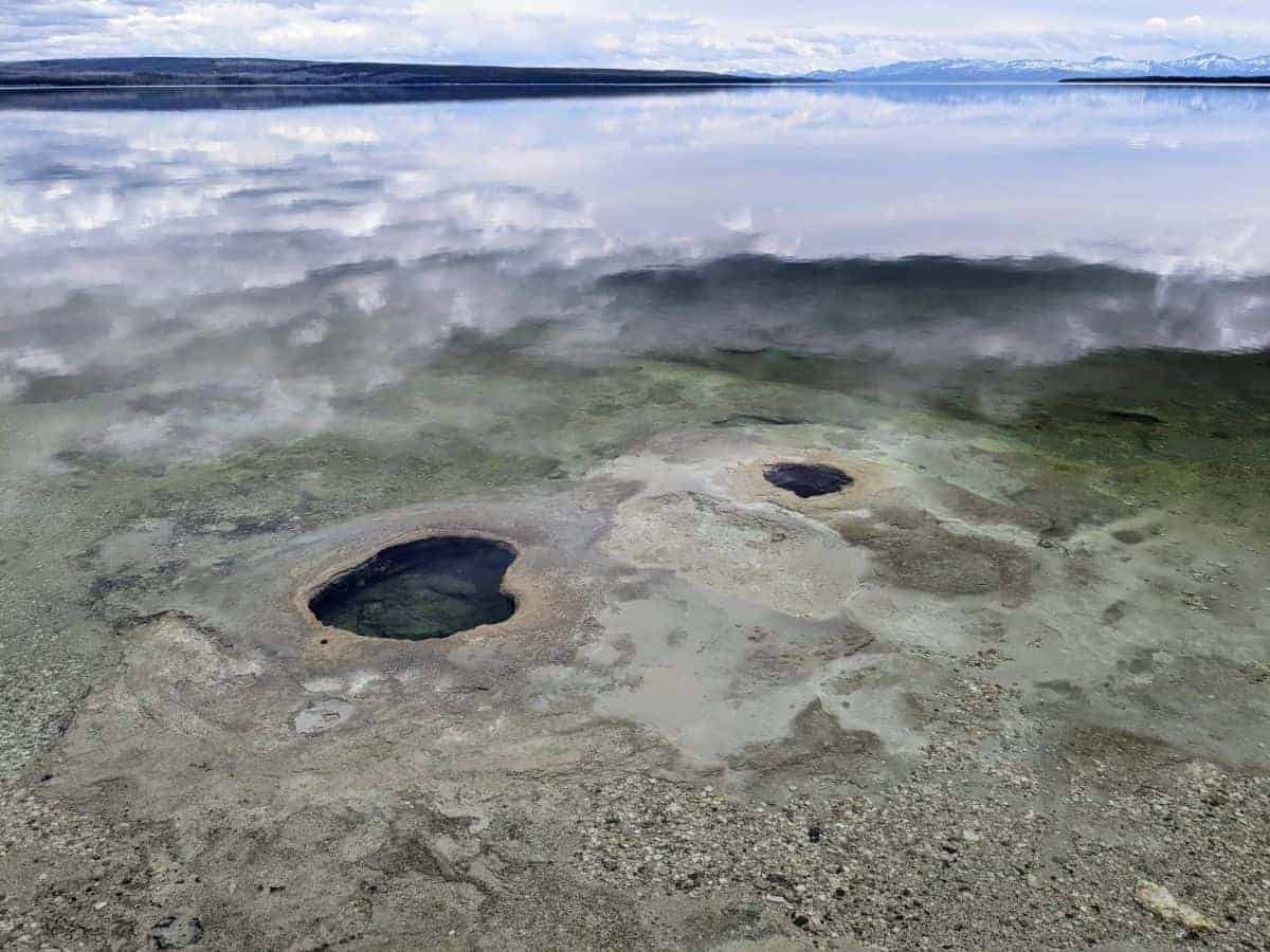 Two holes in the ground under the water on the edge of a lake. There are snowy mountains in the far distance across the lake.