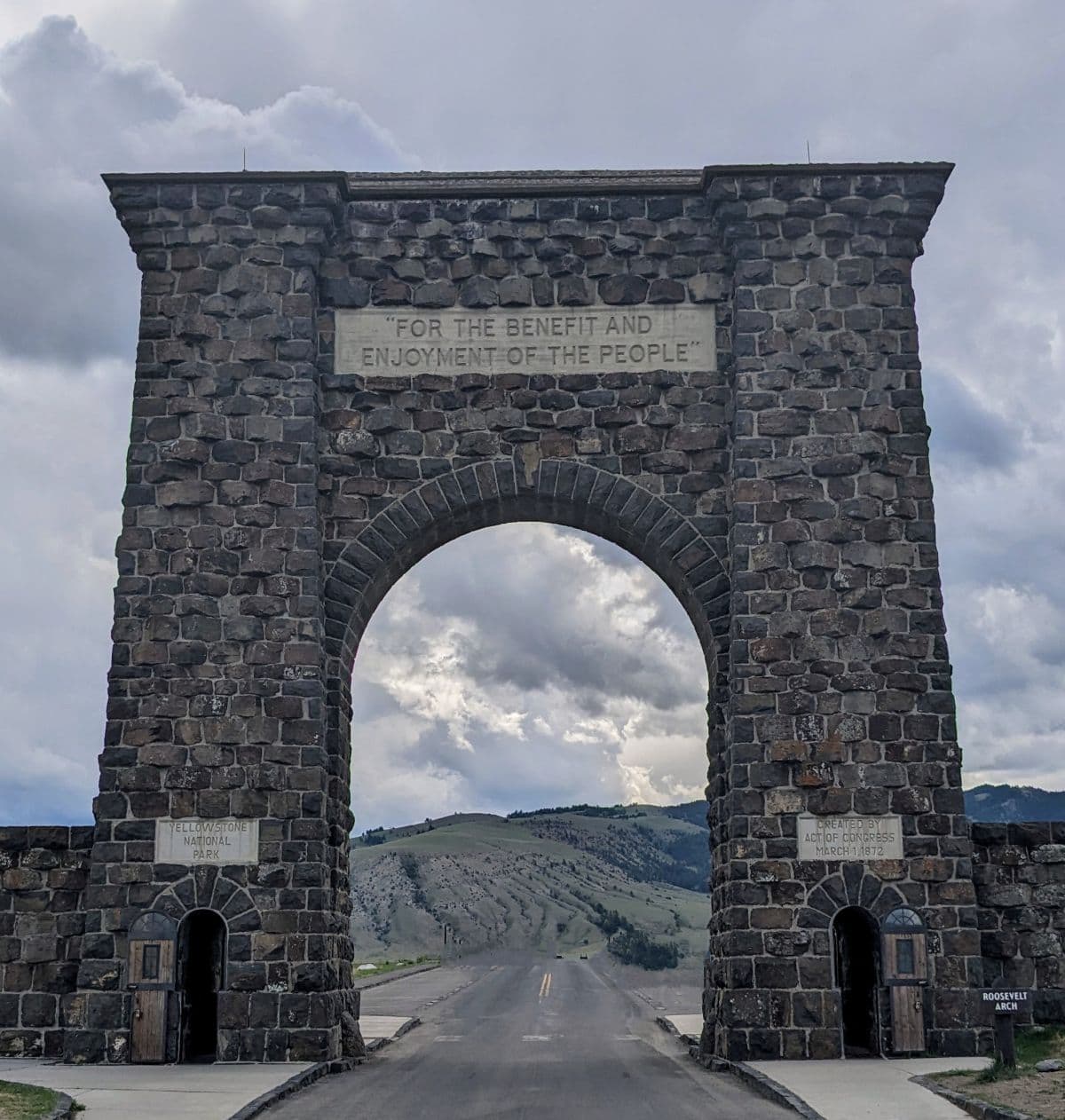 Large stone arch over a roadway with a green hill in the background. At the top of the stone arch there is a chiseled sign that says "For the benefit and enjoyment of the people."