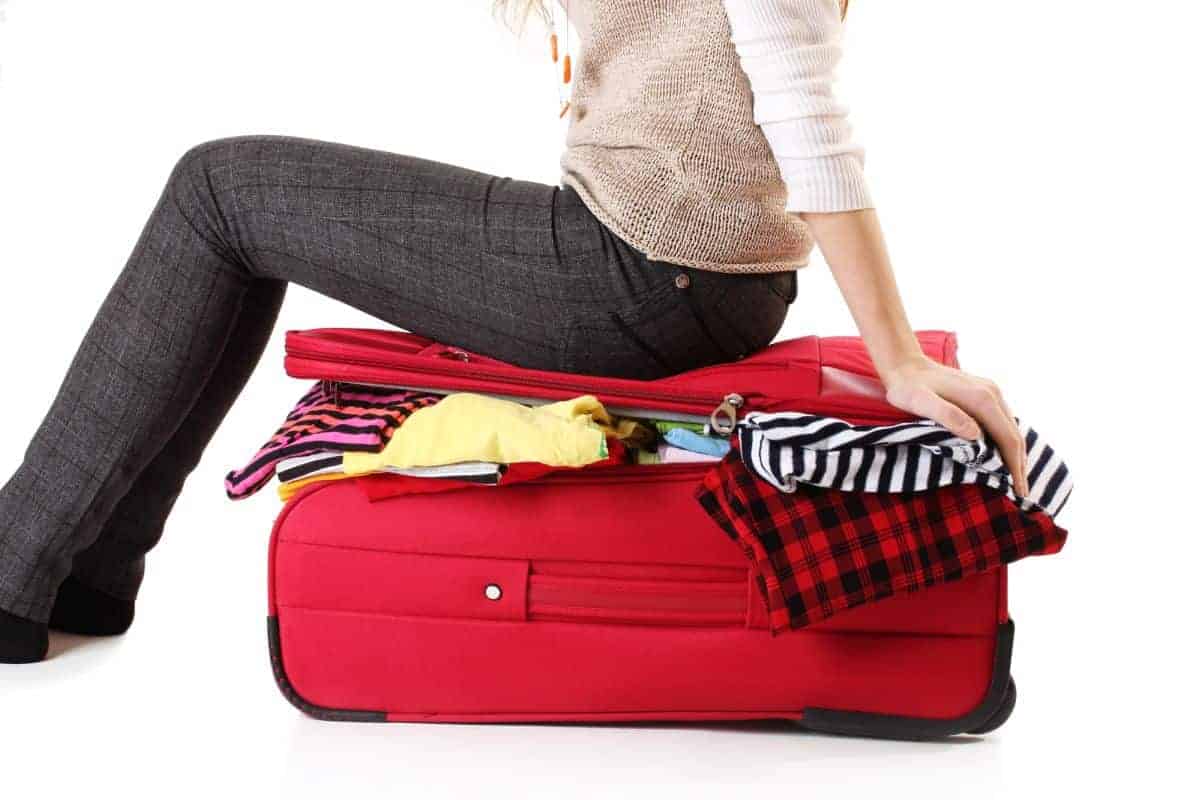 Woman wearing dark grey trousers sitting on a red overstuffed suitcase. Clothing is spilling out.