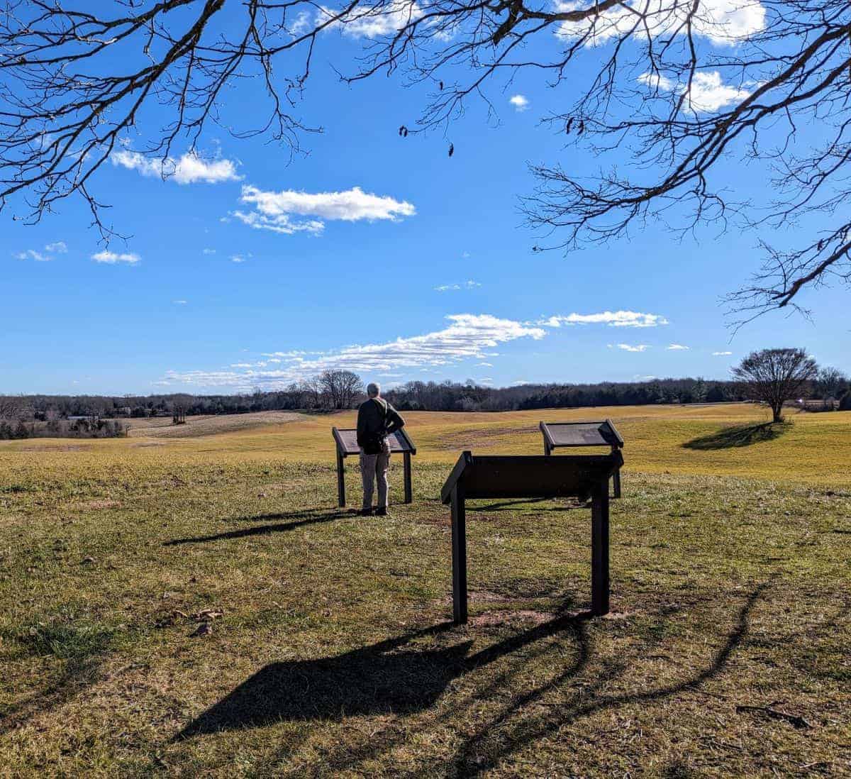 An man stands in front of interpretive signage on a rise overlooking a large open area in Chancellorsville, Virginia.