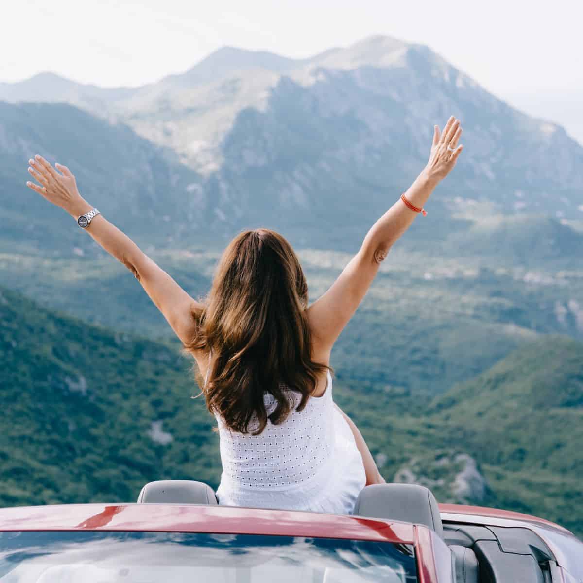 Woman with long dark hair and a white sleeveless outfit sitting on the back of a convertible sportscar. Her arms are raised in happiness as she looks a a lovely mountain view.