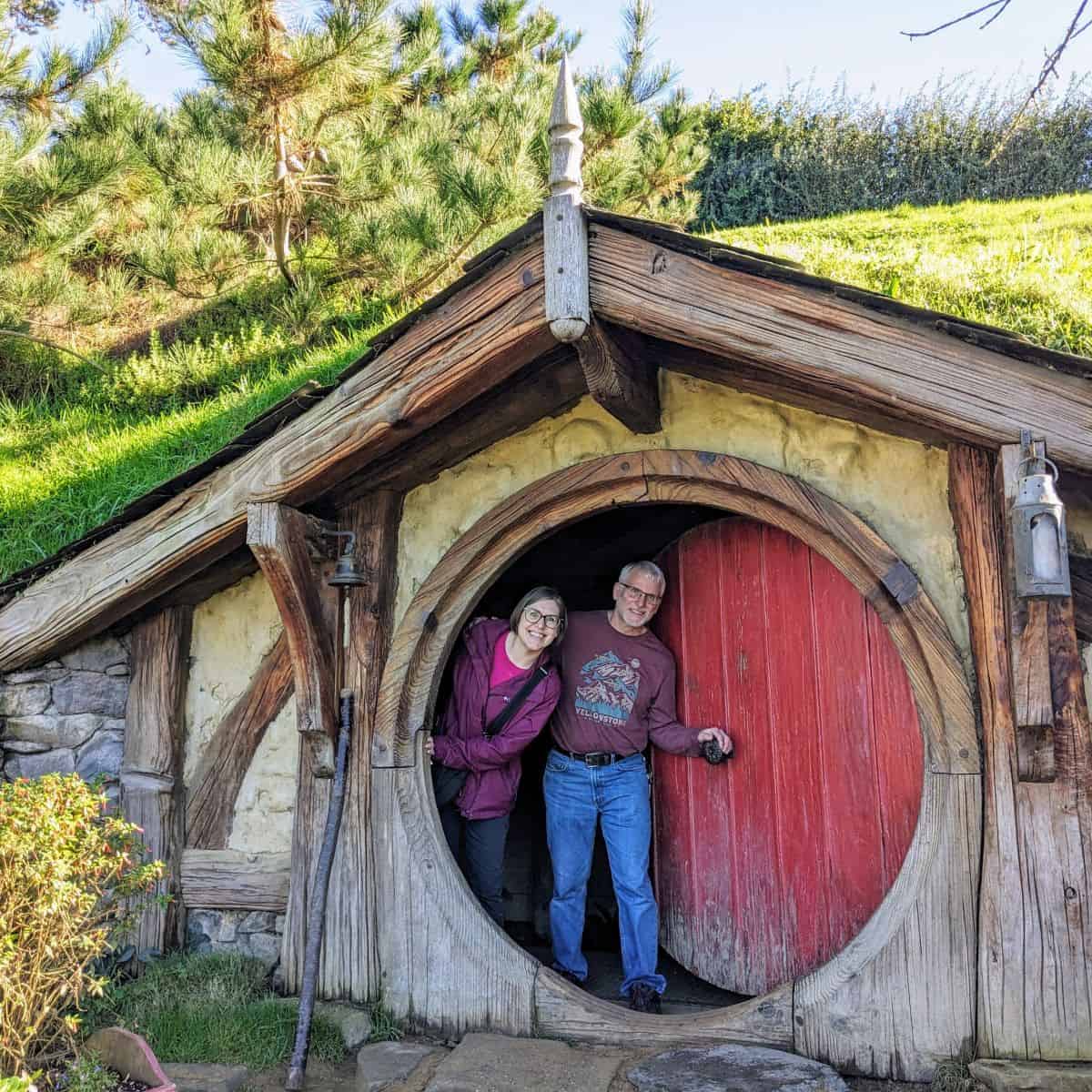 Two people peeking from behind a round red door in a hobbit hole in New Zealand's Hobbiton movie set.