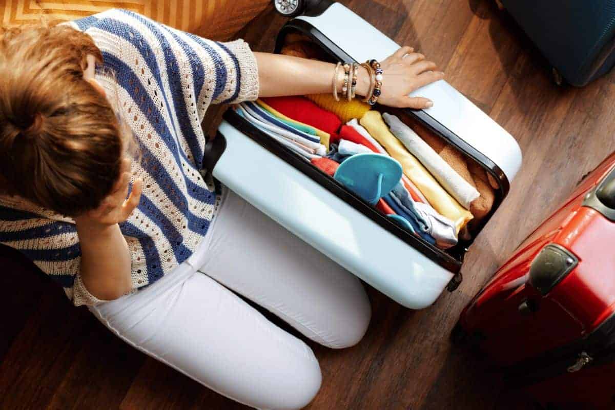 Woman kneeling on a wood floor next to a partly open suitcase that is too full to close. She has her hand on her head like she is stressed about how she is going to manage to get her suitcase closed.
